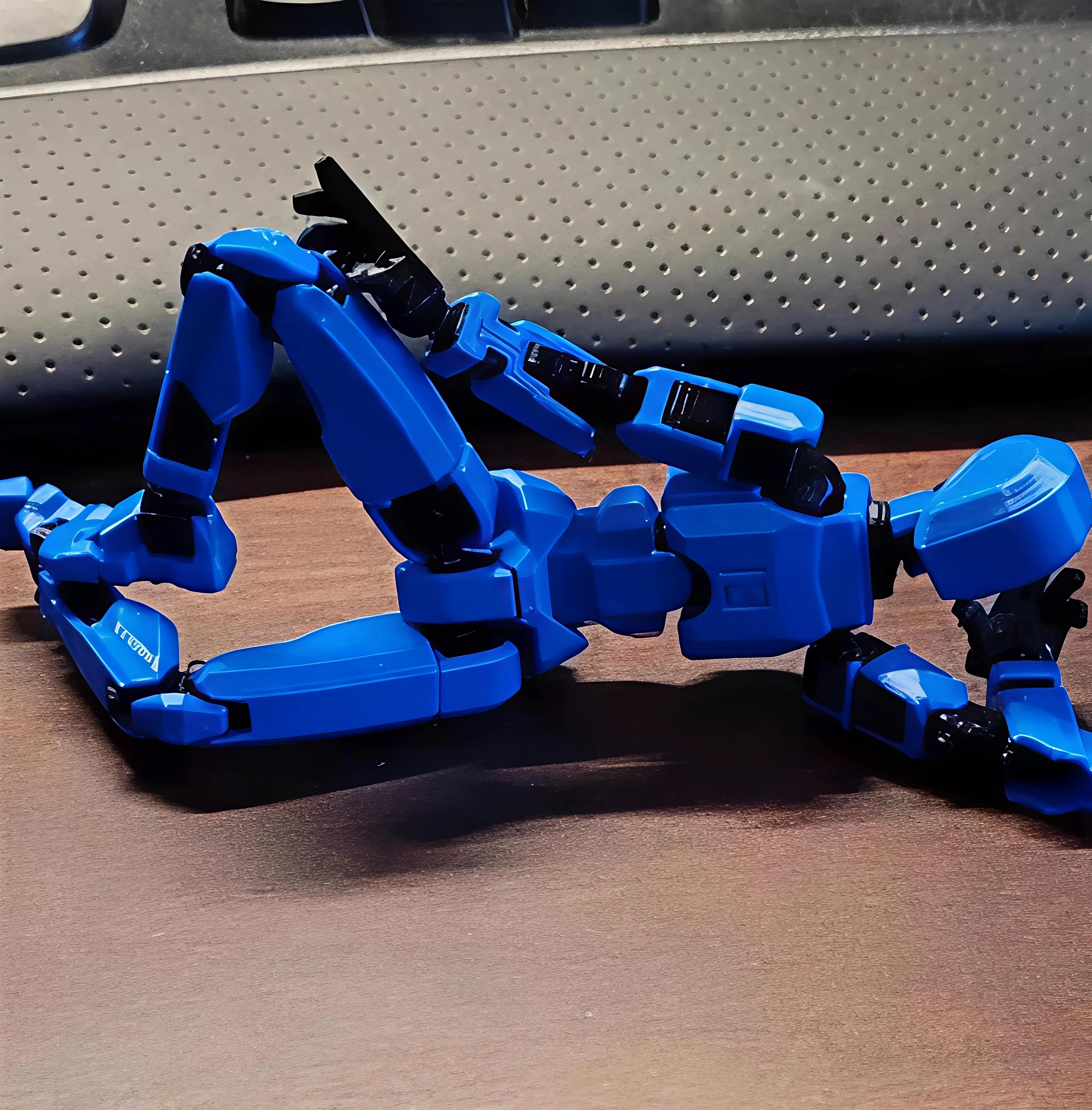 A blue and black articulated robot toy figure posed on a brown wooden surface in front of a gray perforated background.