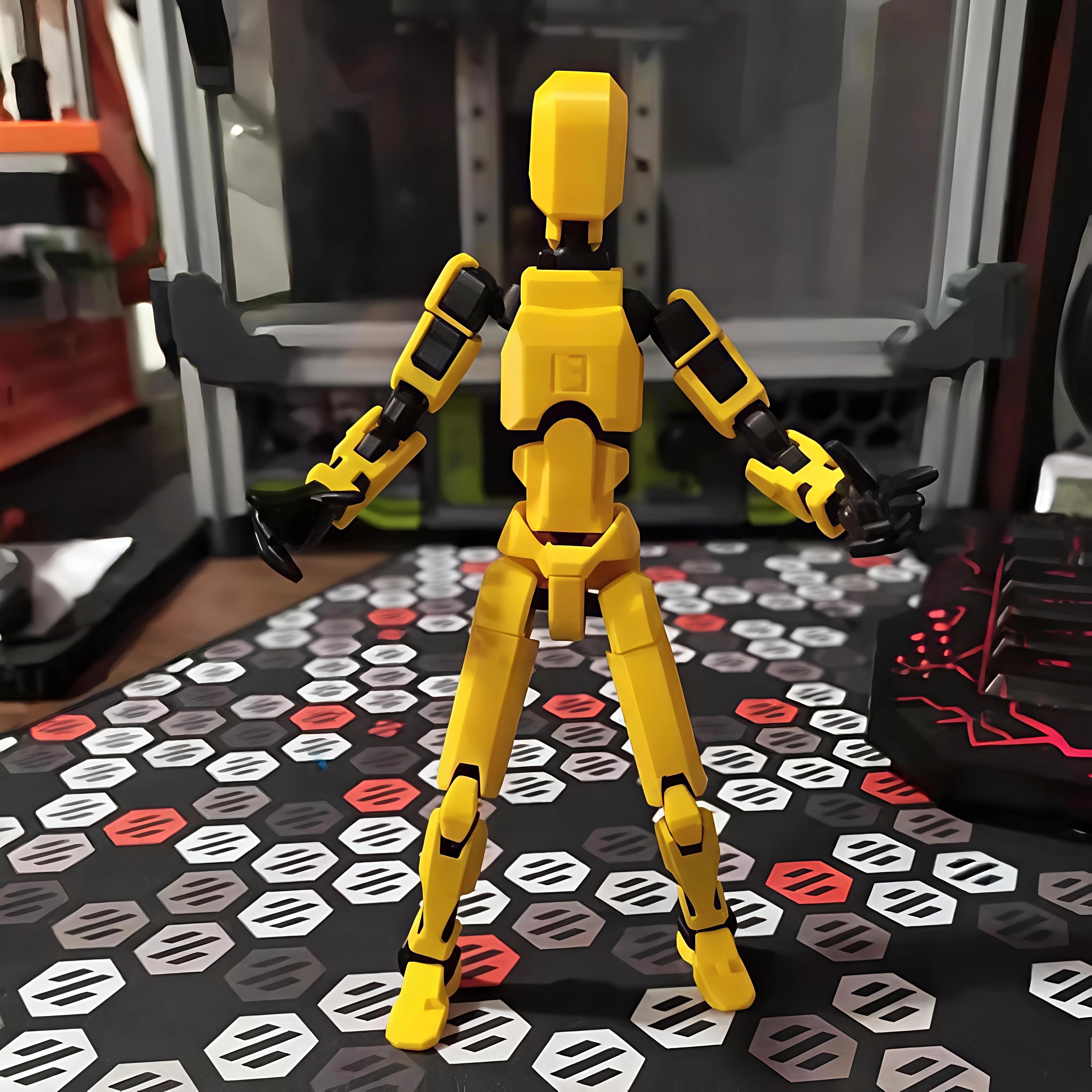 A yellow and black articulated humanoid robot action figure or model stands centered on a patterned desk mat in a desk setting.