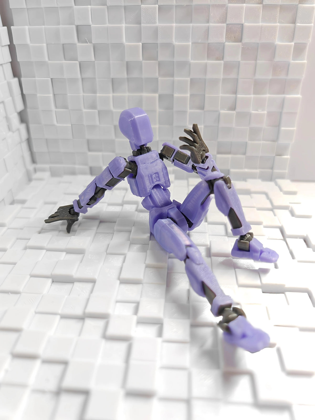 A small purple articulated robot or toy figure with dark gray joints and hands, posed sitting on a white tiled floor against a matching white tiled wall.