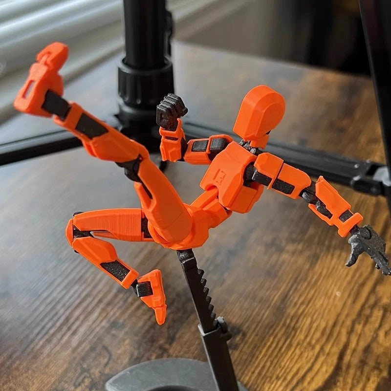 An orange 3D-printed human figure model posed dynamically on a black stand, set against a blurred wooden tabletop background.