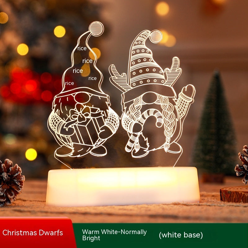 Illuminated Christmas LED lamps with a 'Merry Christmas' sign featuring reindeer antlers and a Christmas tree with presents underneath. The lamps glow warmly, set against a backdrop of a decorated Christmas tree with lights and ornaments. Pine branches and a pine cone add to the festive ambiance.