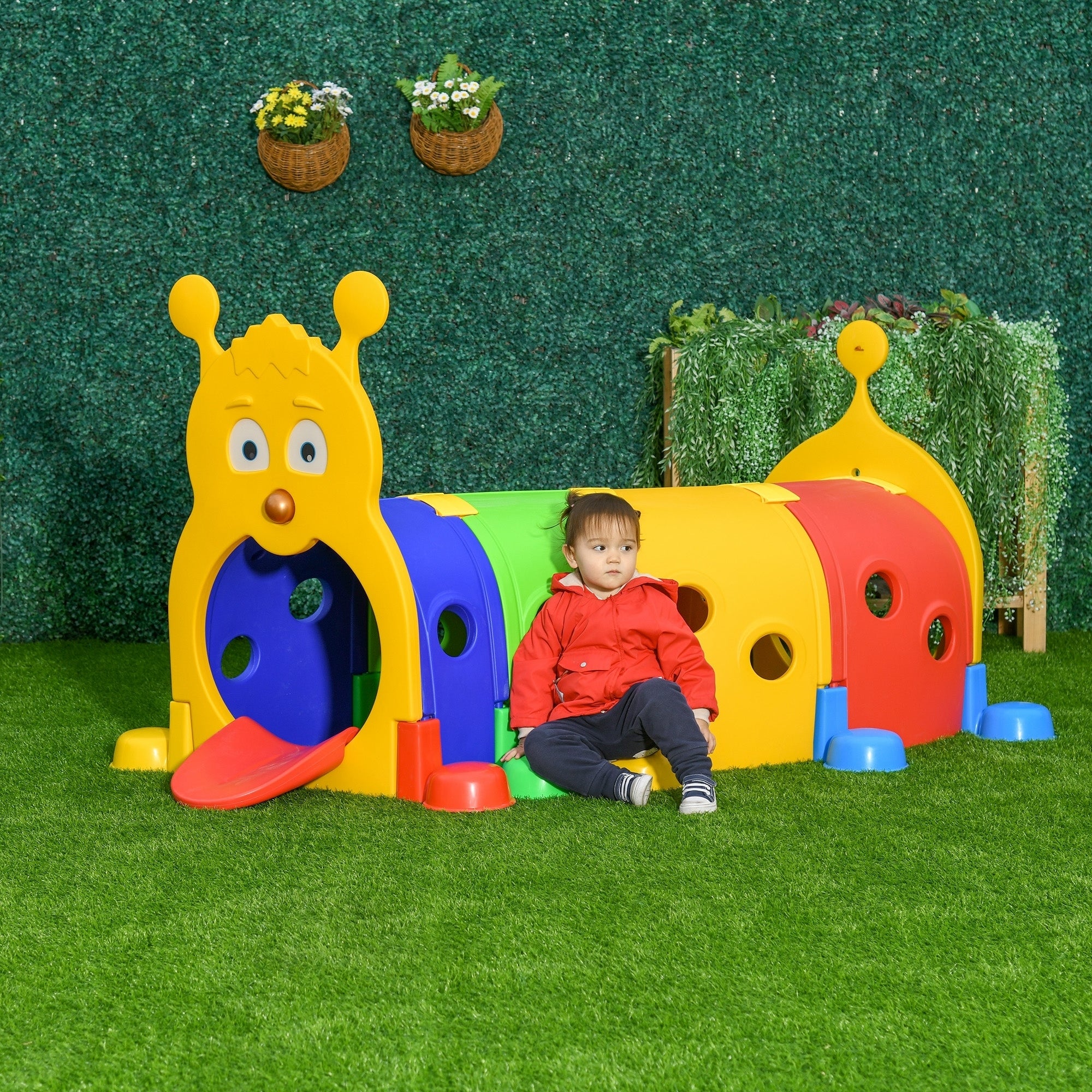 Toddler sitting next to the colorful caterpillar play tunnel in an outdoor garden setting with green grass and a hedge background, highlighting its weather-resistant design.