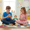 Two children sitting on a colorful classroom carpet, playing with rotating math puzzle toys featuring numbers and symbols. Educational posters and shelves with toys are visible in the background. Toy available at Giggly Groves.